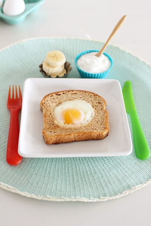 Eggs in a basket on a white plate with a bowl of yogurt and bananas