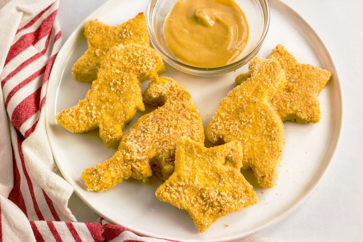 homemade chicken nuggets in dinosaur and star shapes on a white plate with dipping sauce