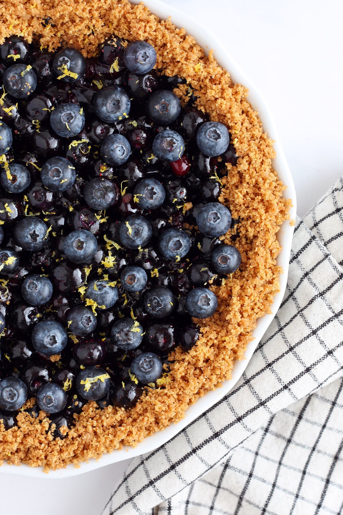 A close up photo of fresh blueberry pie in a graham cracker crust with a checkered linen in the background.