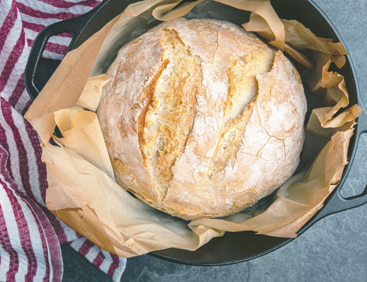 Easy Artisan bread baked in a cast iron dutch oven, just out of the oven, placed on a slate countertop with a kitchen towel waiting to cool