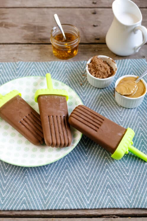 Homemade Peanut Butter Fudgesicle Recipe 1 three peanut butter fudgesicles on a plate with cocoa powder peanut butter and honey in the background