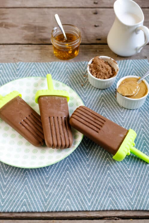 three peanut butter fudgesicles on a plate with cocoa powder peanut butter and honey in the background