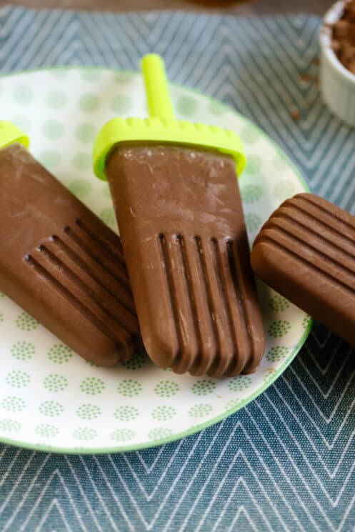 three peanut butter fudgesicles on a plate with cocoa powder peanut butter and honey in the background