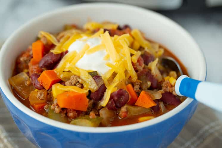 blue bowl of beef and veggie chili with grated cheddar cheese and sour cream on top