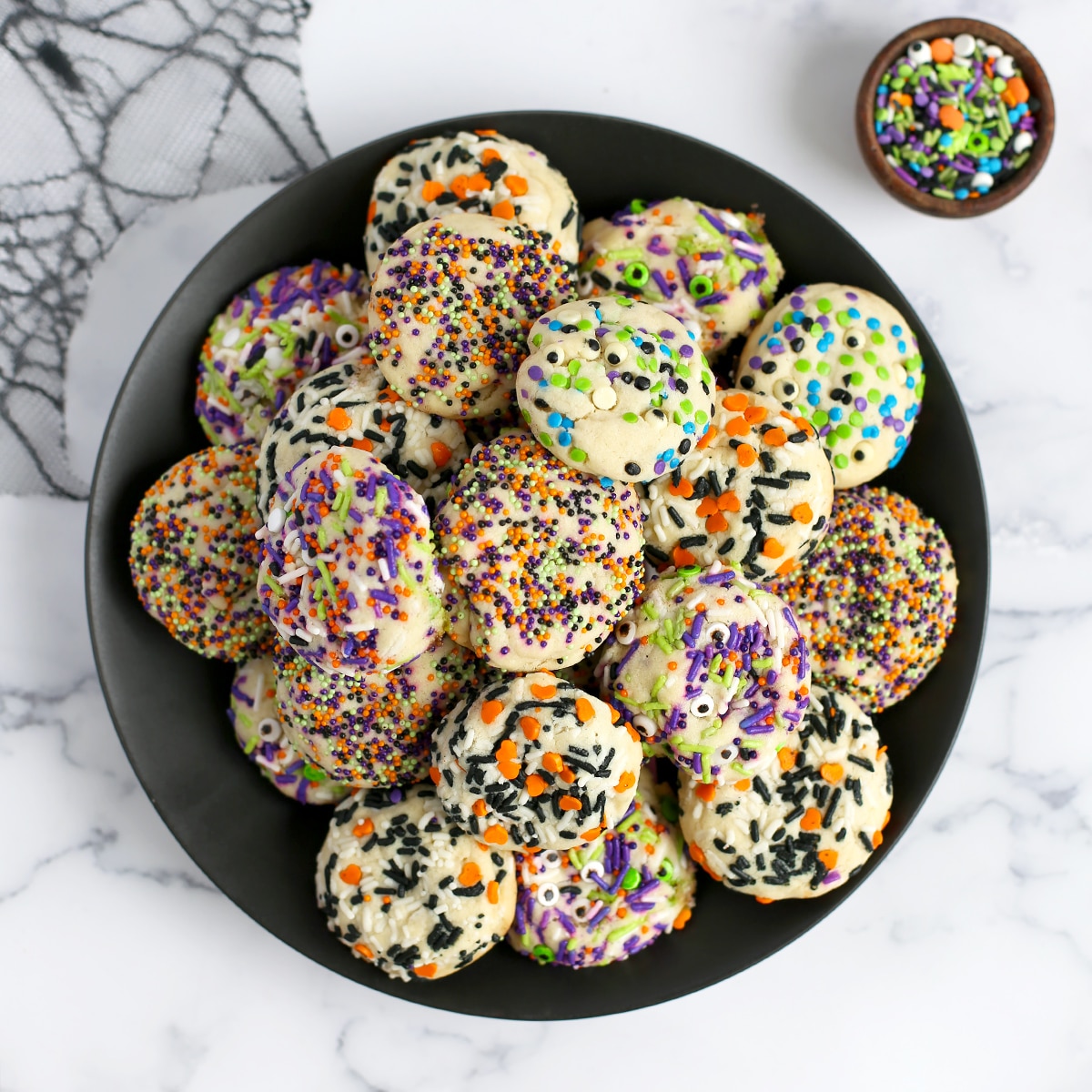 halloween sugar cookies on a black plate with a bowl of sprinkles and spider web in the background