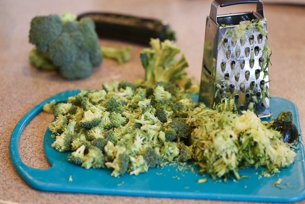 a cutting board and a grater with shredded zucchini and chopped broccoli.