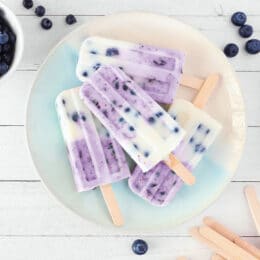 Plate of homemade blueberry vanilla yogurt popsicles. Top view corner border on a white wood background with copy space.