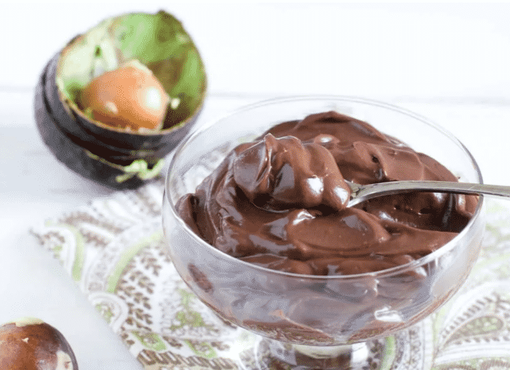 chocolate pudding in a glass bowl with a spoon, with avocado peels and a pit in the background