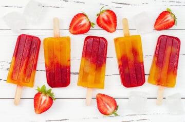 hydrating strawberry mango popsicles in a line with fresh strawberries, and ice on a white wood board