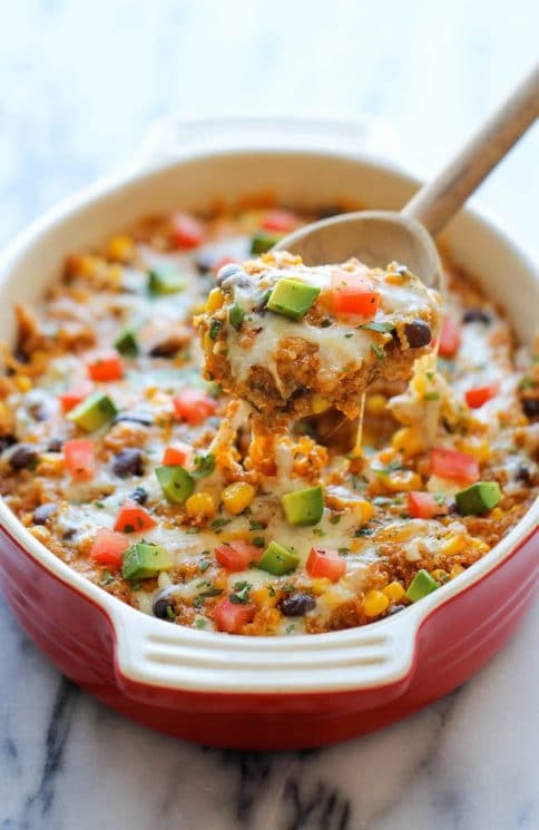 quinoa casserole topped with avocados and tomatoes in a serving dish with a large wooden serving spoon 