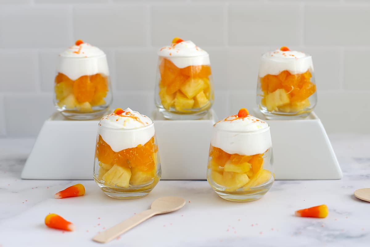 Rows of glass parfait cups with candy corn colored fruit and small wooden spoons.