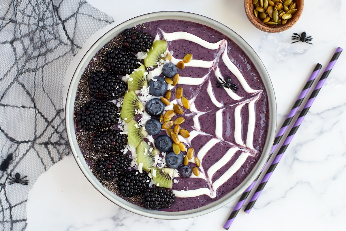 A blueberry smoothie bowl with fruit and nut toppings with purple and black striped straws in the background.