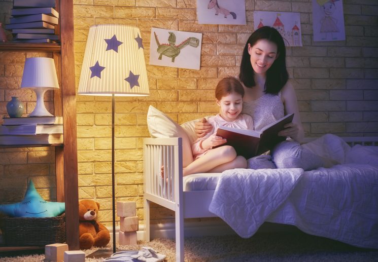 Mom reading a bedtime story to daughter on her bed with a lamp on