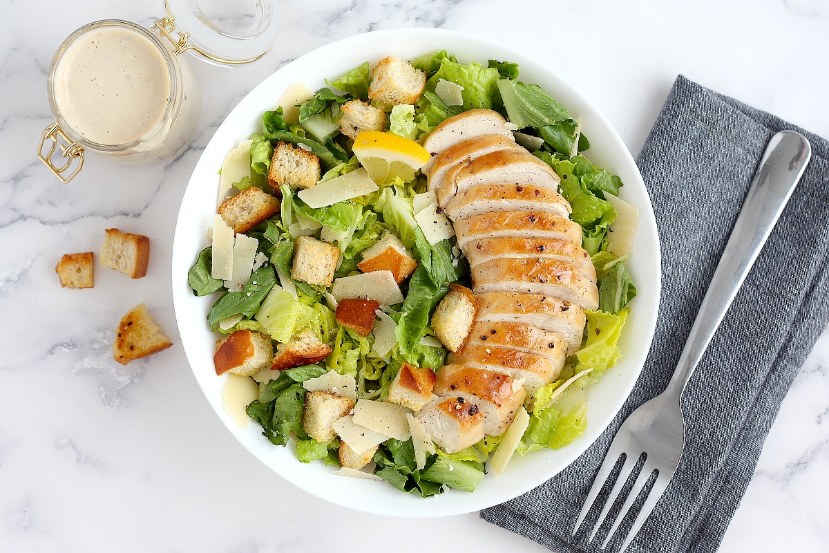 Chicken caesar salad with homemade croutons with a bottle of dressing, a blue linen and a fork.