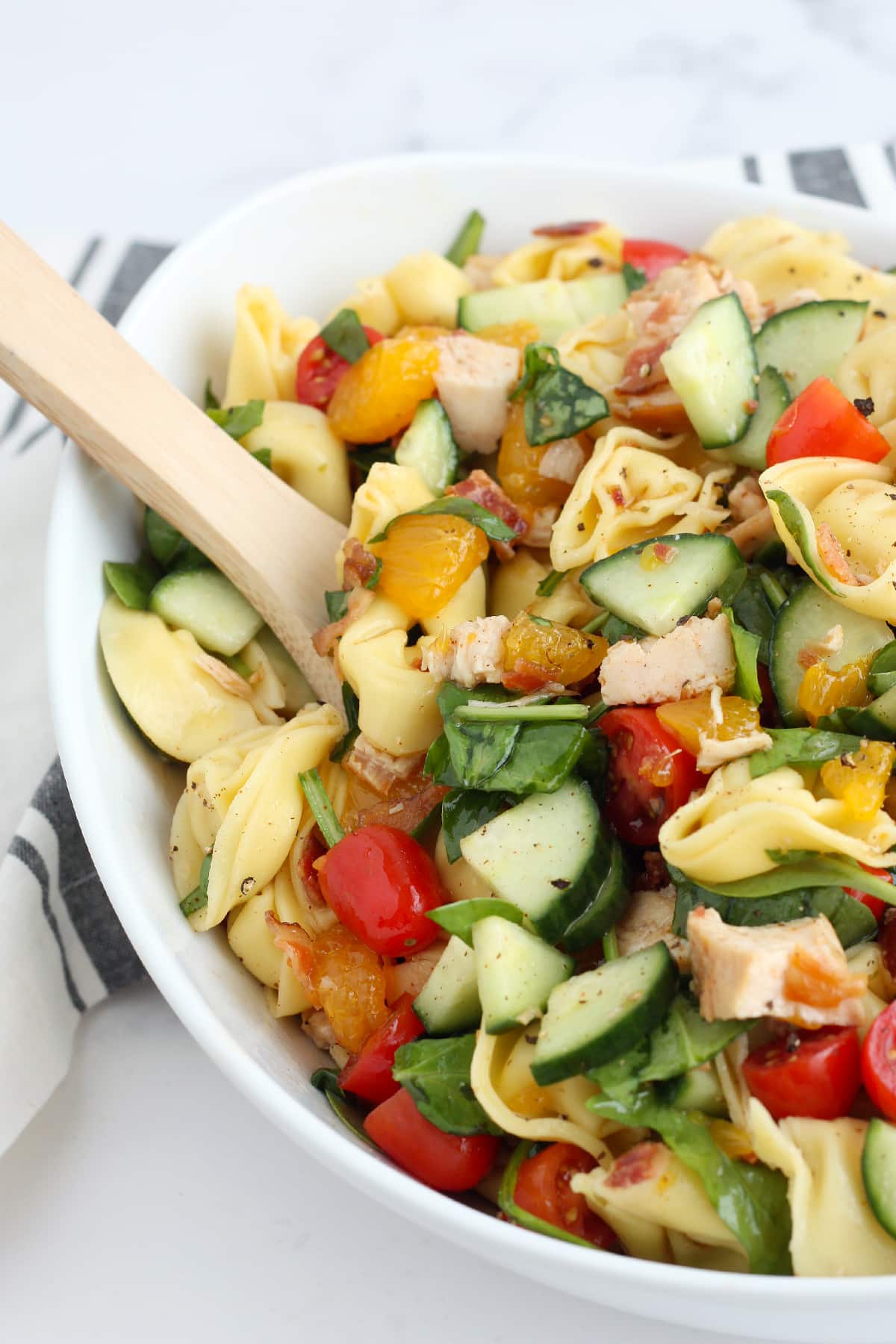 Tortellini pasta salad with chicken, oranges and cucumbers in a white serving bowl with a wooden spoon.