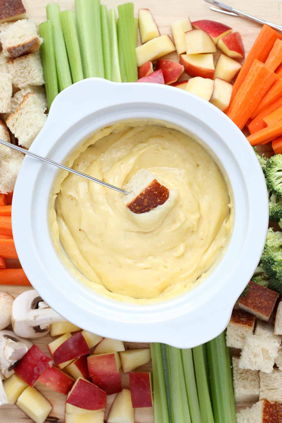 cheese fondue in a white pot with assorted dippers and a bread chunk on a skewer