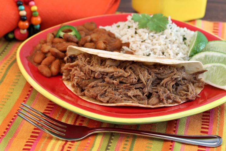 shredded pork soft tacos on a plate with beans and rice and lime wedges