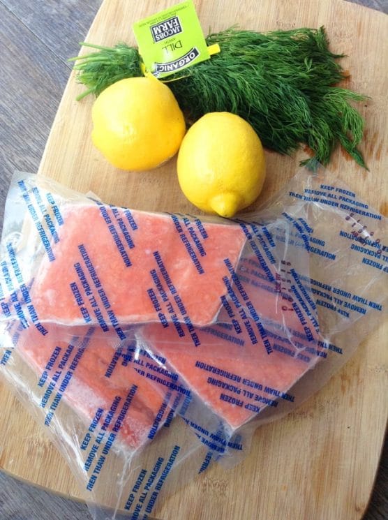 ingredients ready for prep: frozen salmon in bags, whole lemons, and a bunch of dill on a cutting board