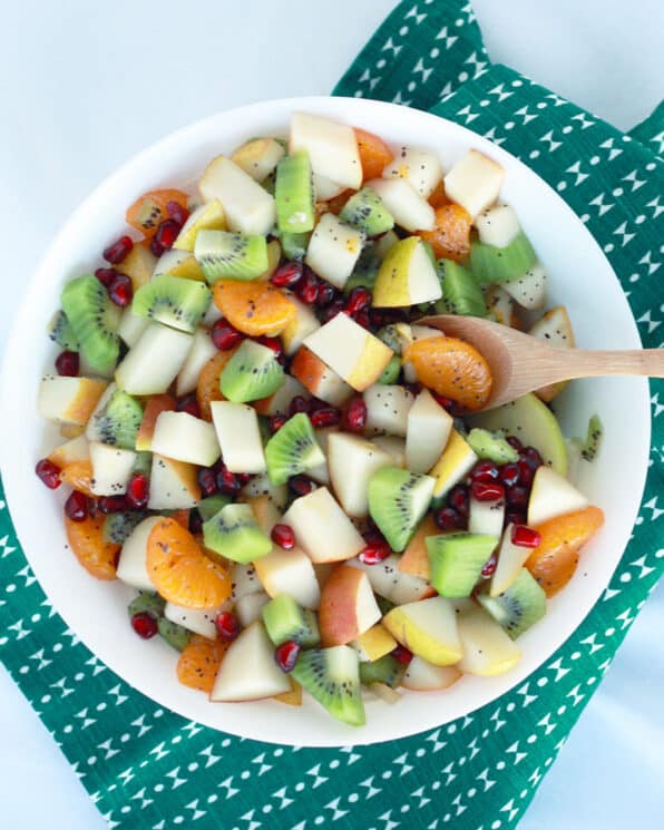 fruit salad made with apples, oranges, kiwi, pear and oranges in a white bowl with a green tea towel in the background