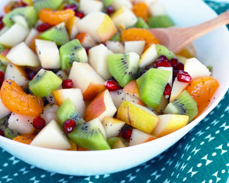 fruit salad made with apples, oranges, kiwi, pear and oranges in a white bowl with a green tea towel in the background