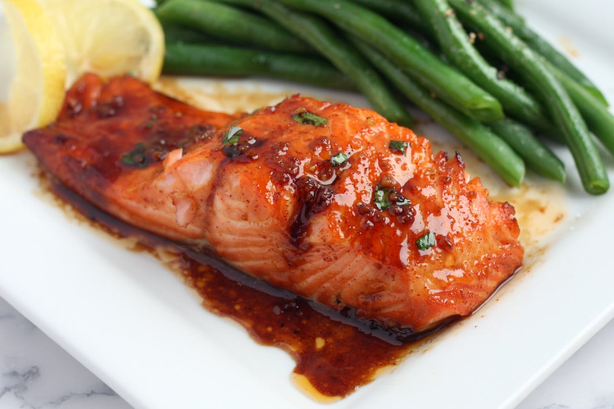 honey garlic salmon on a white square plate with green beans and a lemon slice