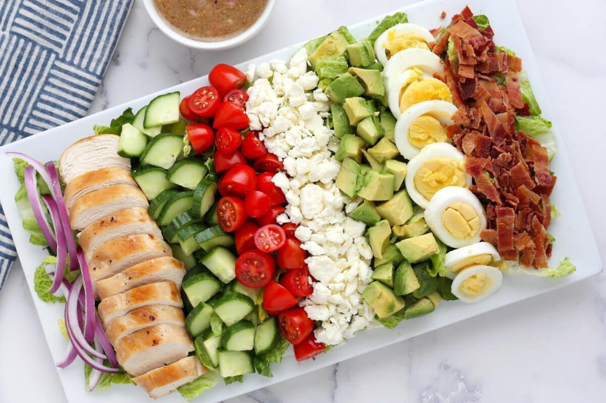 Cobb salad with grilled chicken, hard boiled eggs, crumbled bacon and mixed veggies on a rectangular platter.