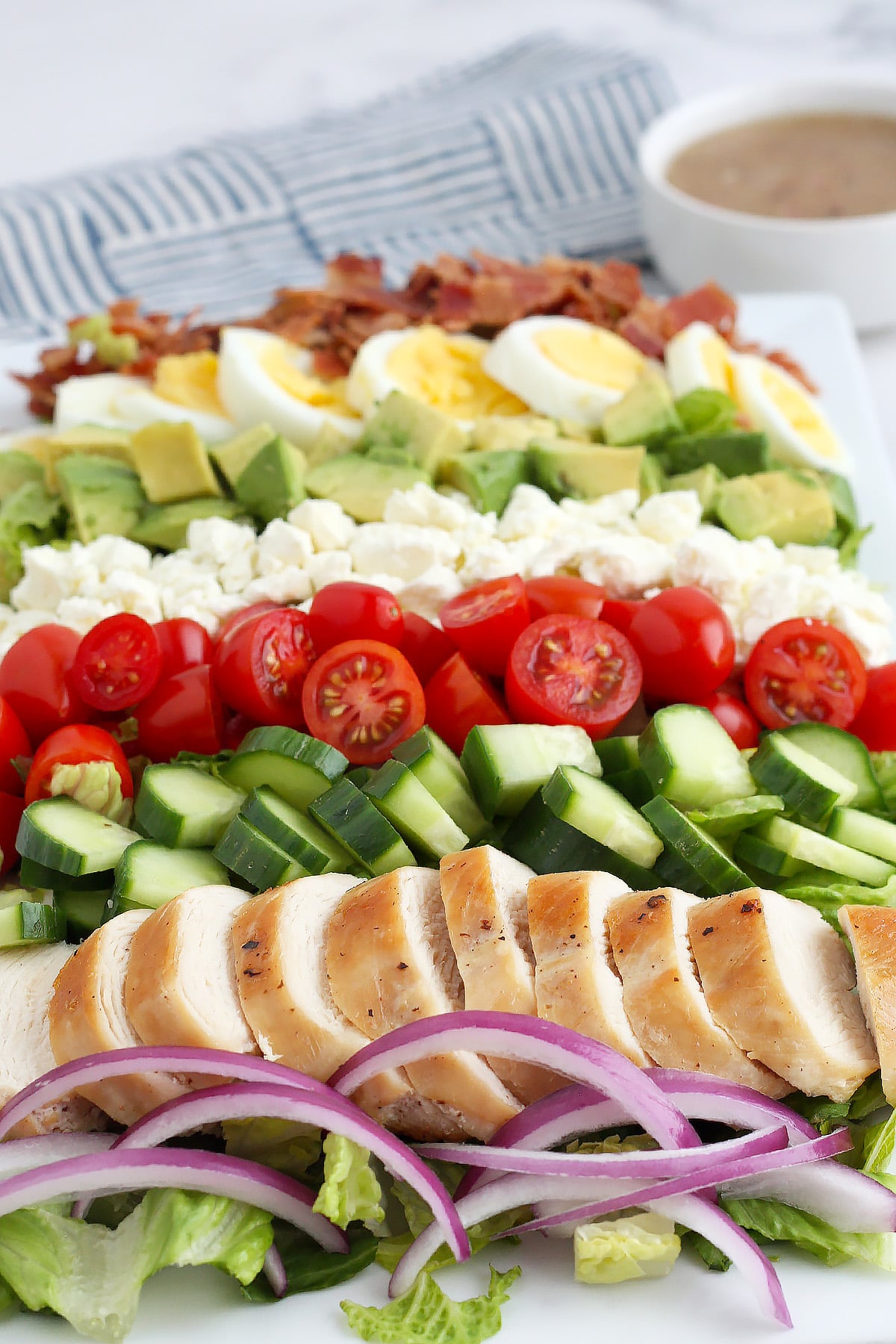 A close up photo of cobb salad with grilled chicken, cucumbers, tomatoes, feta cheese, diced avocado and bacon crumbles.