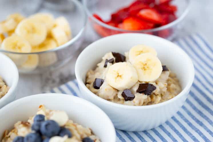 white bowls full of oatmeal with different toppings like bananas, chocolate chips, and blueberries