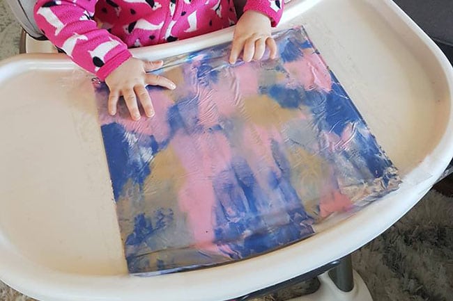 toddler using a plastic bag to smear paint on a high chair