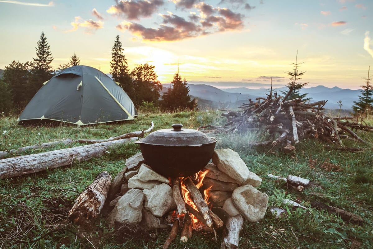 Camp cooking scene with a pot of food cooking on a fire and a tent in the background.