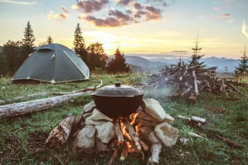 Camp cooking scene with a pot of food cooking on a fire and a tent in the background.