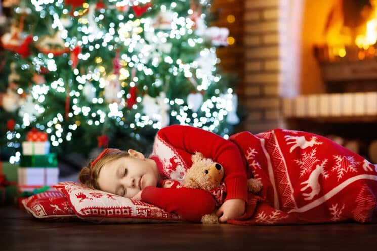 Child sleeping at fire place on Christmas eve under decorated tree. Family celebrating Christmas at home. Kids sleep. Presents at fire place. Little girl under blanket in winter holidays pajamas.