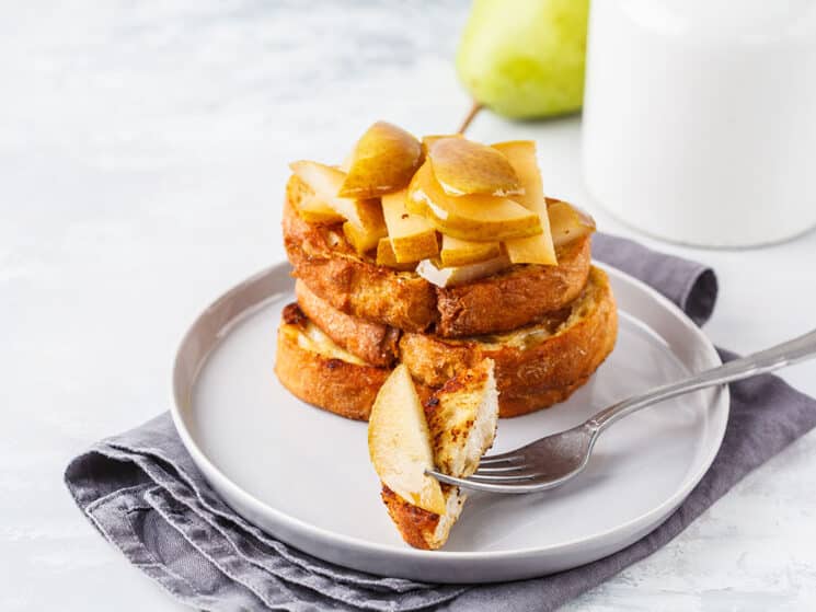 stack of warm pear french toast with a bite cut out on a fork and a pear in the background