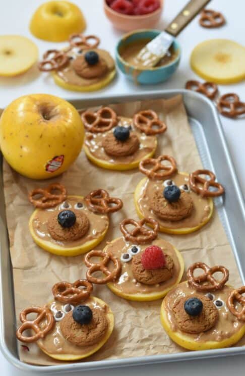 Apple slices topped with peanut butter, cookies and pretzels to resemble reindeer on a baking sheet
