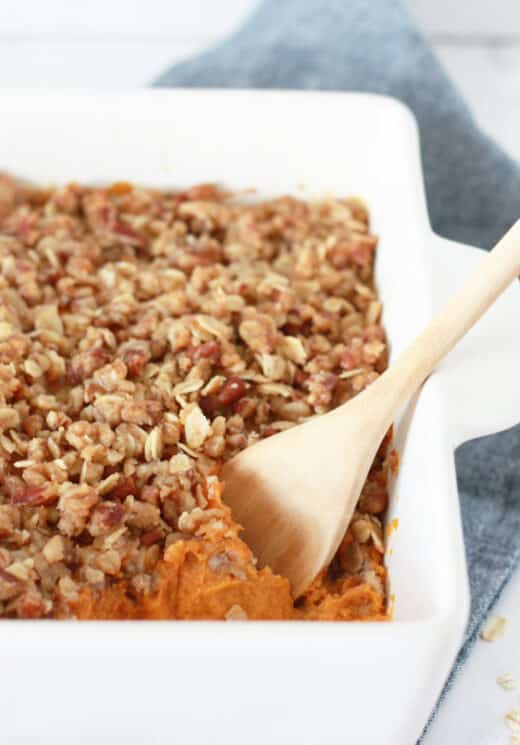 sweet potato casserole with a pecan oat crumble topping in a white baking dish with a wooden serving spoon 