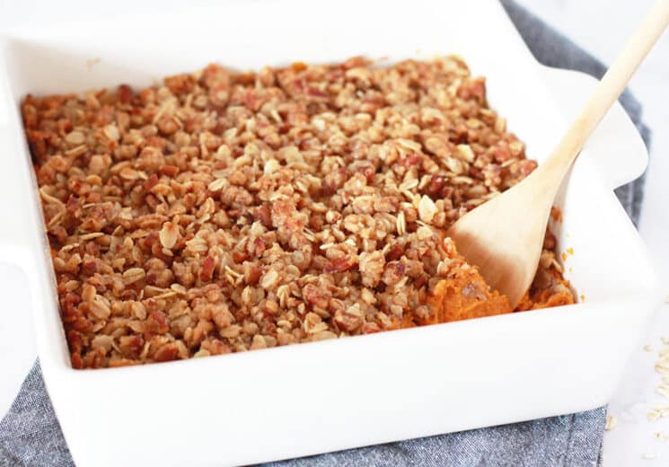 sweet potato casserole with a pecan oat crumble topping in a white baking dish with a wooden serving spoon