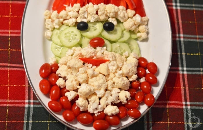 Santa face made with cauliflower, cherry tomatoes, and cucumber slices on a plaid background