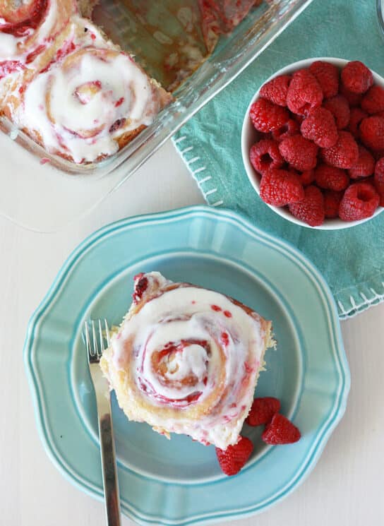 overhead view of a raspberry whole wheat sweet roll on a blue plate with a bowl of fresh raspberries and a pan of the sweet rolls
