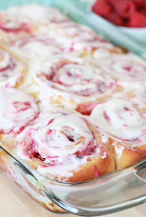 a pan full of raspberry whole wheat sweet rolls with icing on top and fresh raspberries in the background