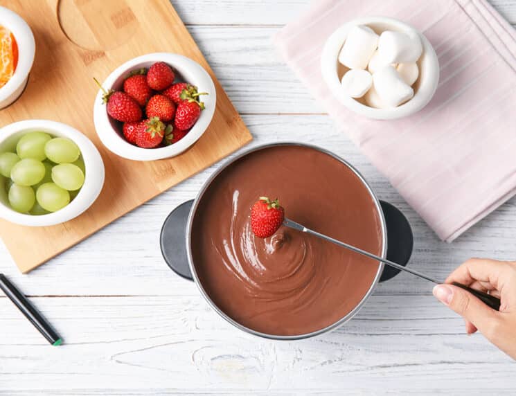 Woman dipping strawberry into pot with chocolate fondue on wooden background, flat lay