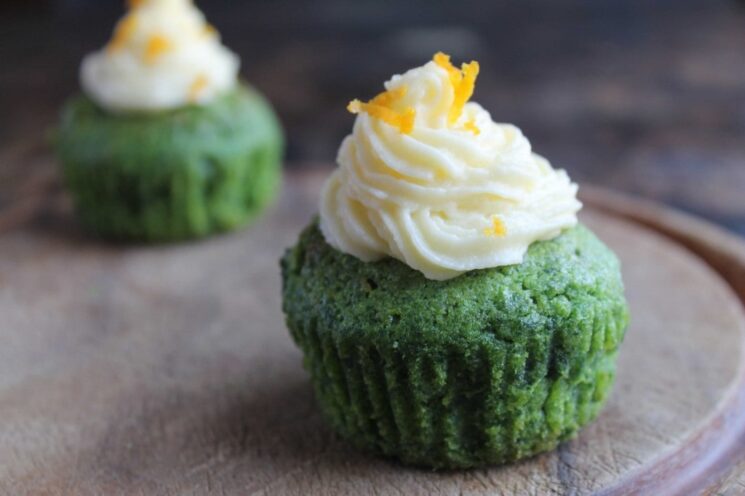 green kale cupcakes topped with a swirl of white frosting and orange zest on a wooden plate