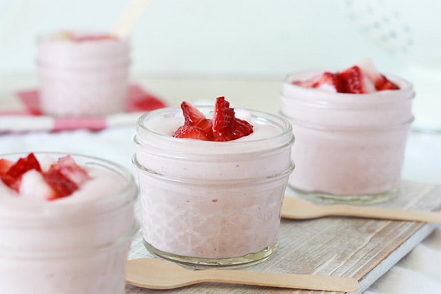 High-protein strawberry mousse in small glass jars with chopped strawberries on top and wooden spoons sit next to jars