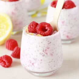 Lemon raspberry chia seed pudding in a small glass jar with fresh raspberries and lemon zest on top, a small silver spoon in the jar and fresh lemons in the background