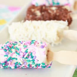 frozen banana pops on a white tray with different toppings: sprinkles, coconut, chocolate pieces