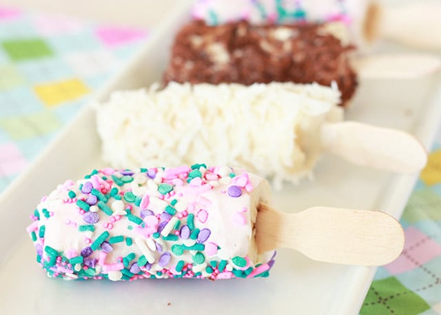 frozen banana pops on a white tray with different toppings: sprinkles, coconut, chocolate pieces