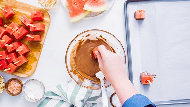 Overlay view of someone dipping watermelon in chocolate and placing it on a parchment-lined baking sheet