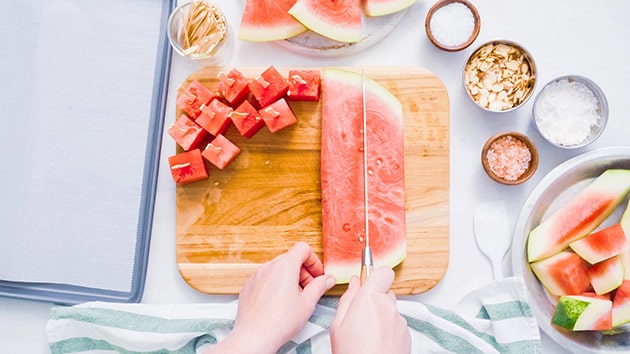 overlay view of cutting watermelon into cubes on a wooden cutting board.  Cubes of watermelon with popsicle sticks sitting on the cutting board.