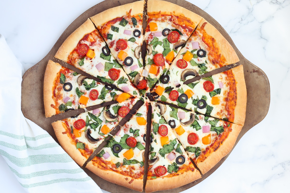 A sliced vegetable pizza on a pizza stone with a green striped linen.