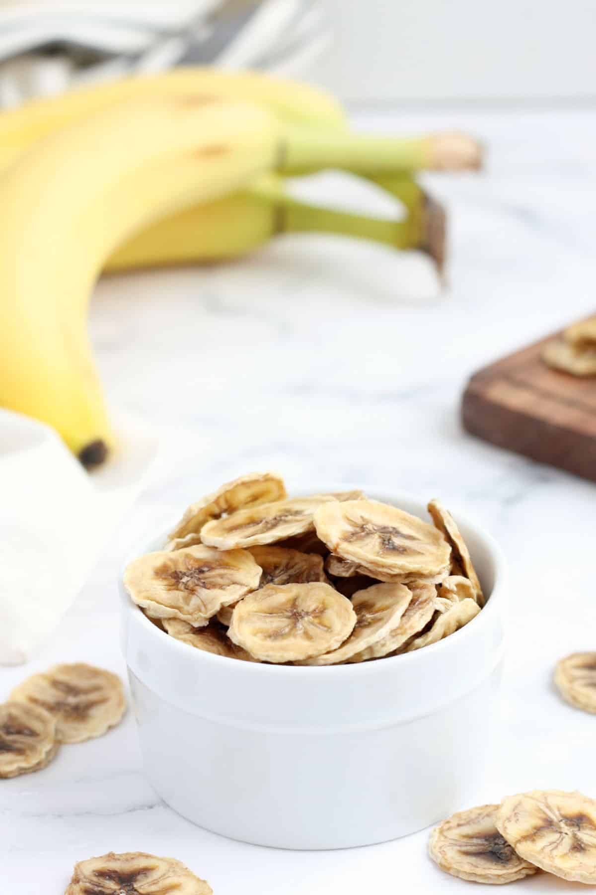 homemade banana chips in a white bowl with fresh bananas in the background
