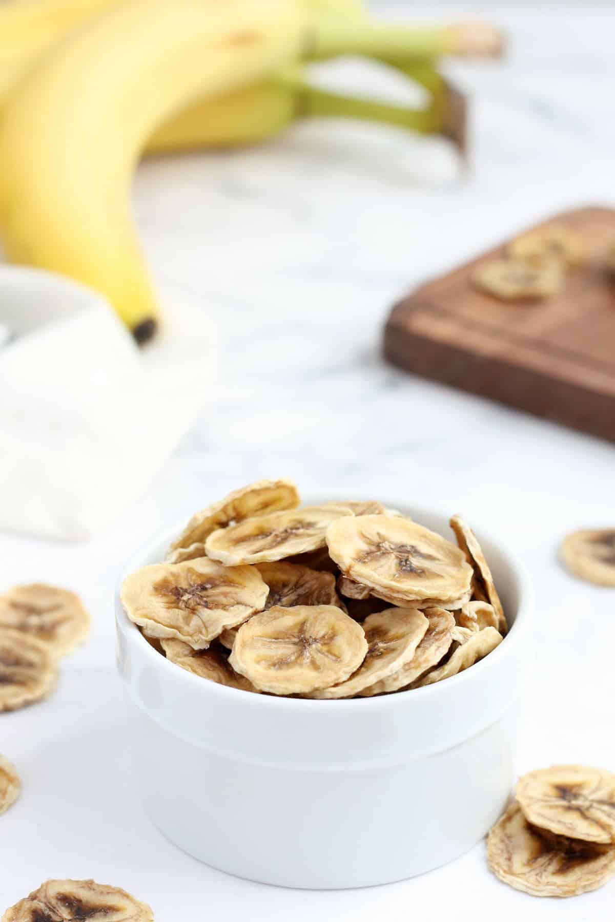 homemade banana chips in a white bowl with fresh banana in the background